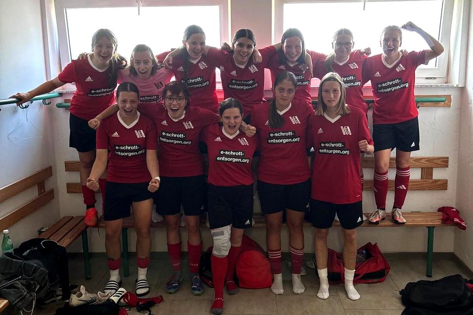 Gruppe von zwölf Fußballspielerinnen in roten Trikots, fröhlich posierend in einer Umkleidekabine.