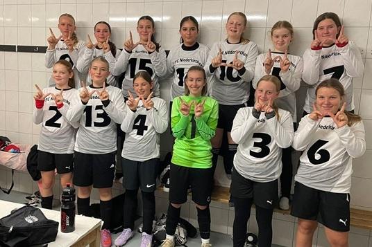 Mannschaftsaufnahme von zwölf Fußballerinnen in Umkleide mit Trikots und Siegerzeichen. Torhüterin trägt grüne Ausrüstung.