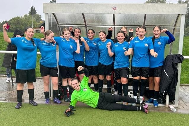 Gruppe von elf Fu&szlig;ballspielerinnen in blauen Trikots, einige jubeln, w&auml;hrend eine Torh&uuml;terin auf dem Boden liegt.