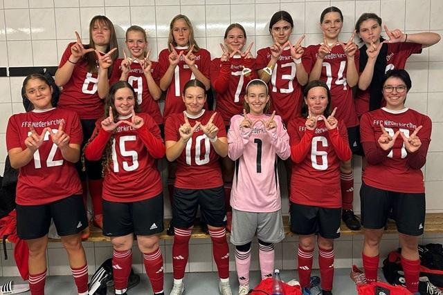 Mannschaftsfoto von zw&ouml;lf Fu&szlig;ballspielerinnen in roten Jerseys mit verschiedenen R&uuml;ckennummern in einem Umkleideraum.