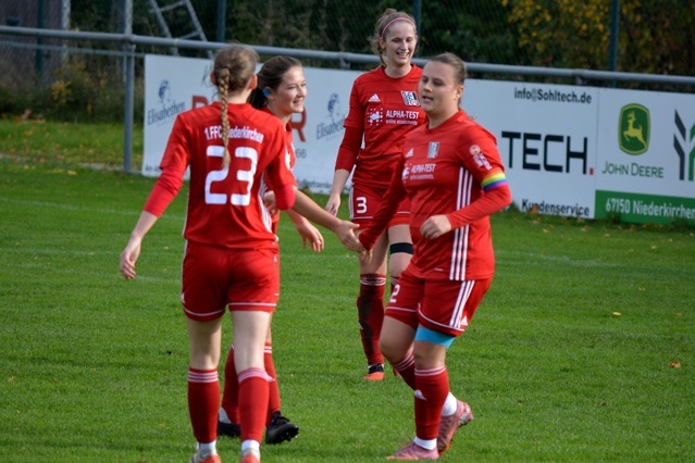 Vier Fu&szlig;ballspielerinnen in roten Trikots feiern auf dem gr&uuml;nen Platz nach einem gelungenen Spielzug.