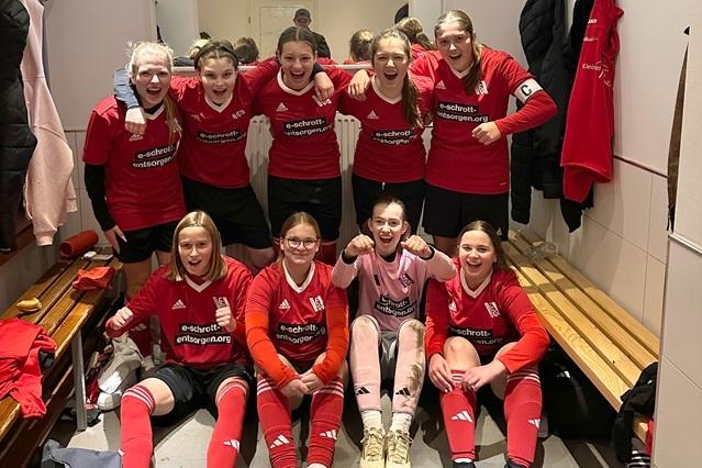 Mannschaftsfoto von elf M&auml;dchen in roten Fu&szlig;balltrikots, l&auml;chelnd und jubelnd in einer Umkleidekabine.