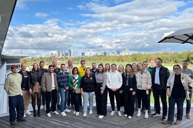 Gruppe von 24 Personen steht l&auml;chelnd im Freien mit Stadtsilhouette und bew&ouml;lktem Himmel im Hintergrund.