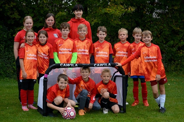 Gruppe von Kindern in orangefarbenen Fu&szlig;balltrikots posiert vor einem Tor, umgeben von gr&uuml;ner Landschaft.