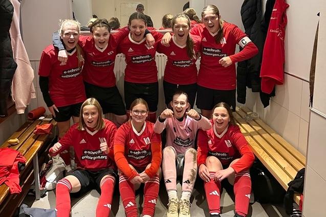 Mannschaftsfoto von elf M&auml;dchen in roten Fu&szlig;balltrikots, l&auml;chelnd und jubelnd in einer Umkleidekabine.