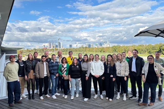 Gruppe von 24 Personen steht lächelnd im Freien mit Stadtsilhouette und bewölktem Himmel im Hintergrund.
