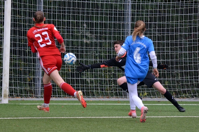 Torwart in schwarzer Ausr&uuml;stung versucht, einen Ball von einer laufenden Spielerin im roten Trikot abzuwehren.