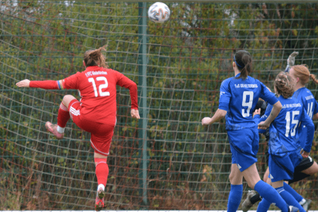 Spielszene mit einer Torwartspielerin in Rot, die einen Ball abwehrt, w&auml;hrend Spielerinnen in Blau zuschauen.