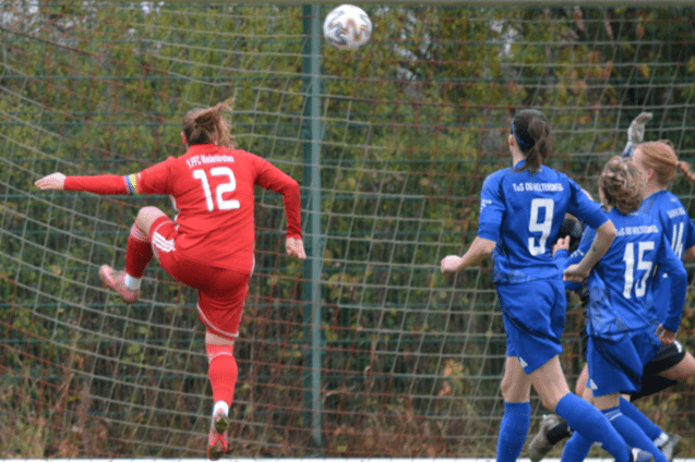 Spielszene mit einer Torwartspielerin in Rot, die einen Ball abwehrt, w&auml;hrend Spielerinnen in Blau zuschauen.