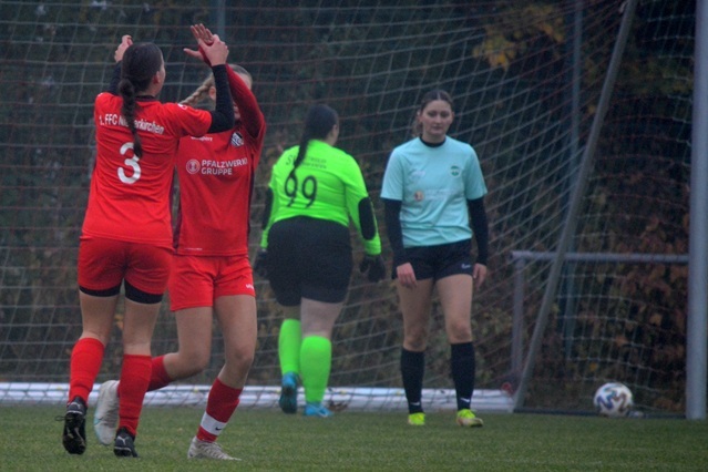 Zwei Fu&szlig;ballspielerinnen in roten Trikots feiern ein Tor, w&auml;hrend eine Torh&uuml;terin in gr&uuml;ner Ausr&uuml;stung im Hintergrund steht.