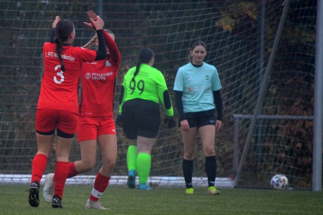 Zwei Fu&szlig;ballspielerinnen in roten Trikots feiern ein Tor, w&auml;hrend eine Torh&uuml;terin in gr&uuml;ner Ausr&uuml;stung im Hintergrund steht.