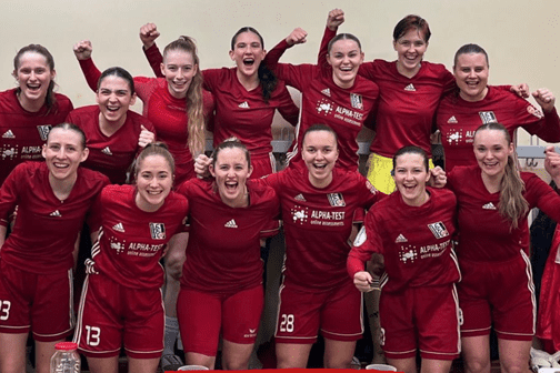 Eine jubelnde Frauenfußballmannschaft in roten Trikots posiert nach einem Sieg von 6-0, Ergebnisinfos im Vordergrund.