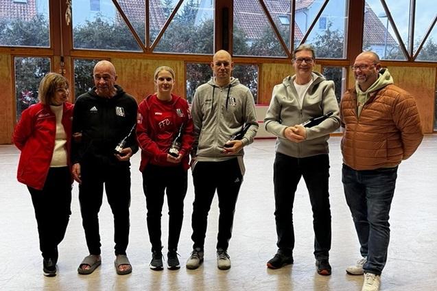 Gruppe von sechs Personen, die Troph&auml;en in einer Sporthalle pr&auml;sentieren, mit gro&szlig;en Fenstern im Hintergrund.