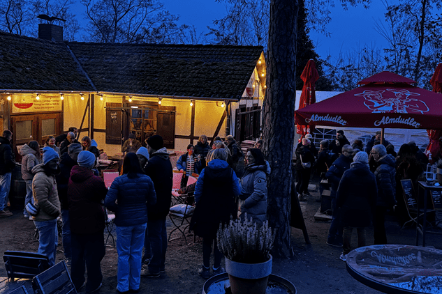 Eine Gruppe von Menschen in warmen Jacken versammelt sich am Abend vor einem beleuchteten Gasthaus im Freien.