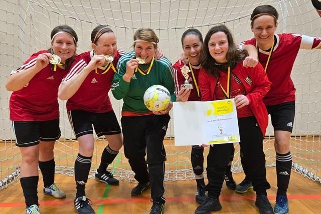 Sechs Frauen im Fußballtrikot posieren mit Medaillen, Pokalen und einer Urkunde vor einem Fußballtor.