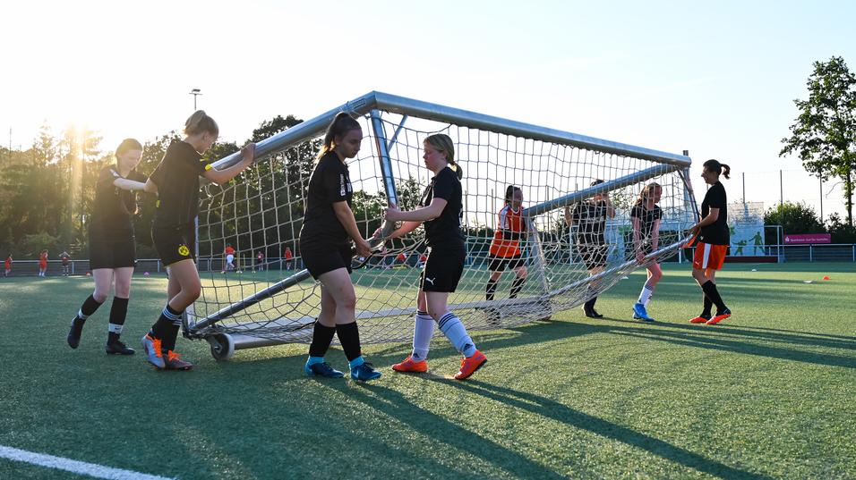 Mannschaft von Fußballspielerinnen hebt ein Tor auf einem Kunstrasenplatz bei Sonnenuntergang an.