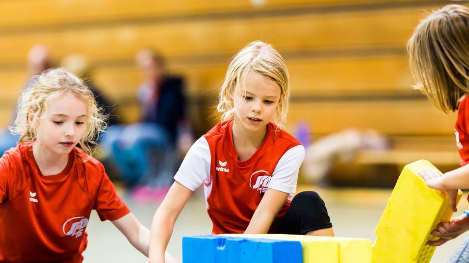 Drei Mädchen im roten Shirt bauen mit bunten Schaumstoffbausteinen in einer Turnhalle.